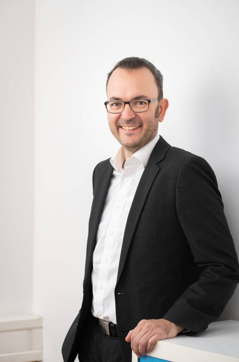 Portraitbild von Prof. Dr. Daniel Pak, der an einem weißen Regal lehnt und in die Kamera lacht.